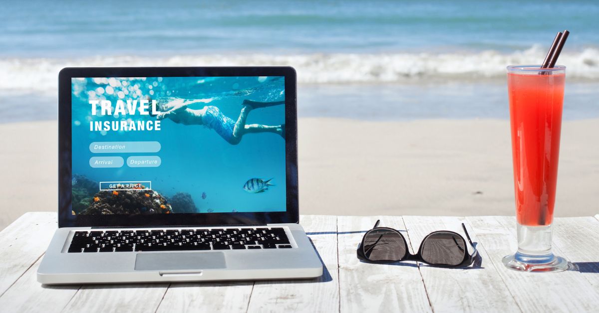 Laptop dengan layar menampilkan halaman web untuk beli asuransi perjalanan online diletakkan di pantai dengan minuman tropis dan kacamata hitam.